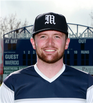 Photograph of Coach Ranieri in blue shirt and baseball cap with an M on the cap. Standing in front of a baseball scoreboard.